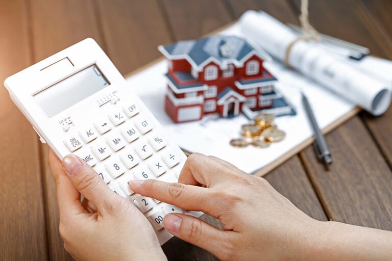 Mujer usa una calculadora mientras al fondo se ve una maqueta de casa sobre unos planos